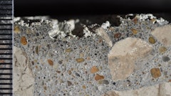 Photograph of polished surface from a concrete core removed from a poorly cured slab on ground showing layer of dark paste at the top of the core. Scale in millimeters. The loss of material at the top is due to poor hydration.