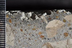 Photograph of polished surface from a concrete core removed from a poorly cured slab on ground showing layer of dark paste at the top of the core. Scale in millimeters. The loss of material at the top is due to poor hydration.