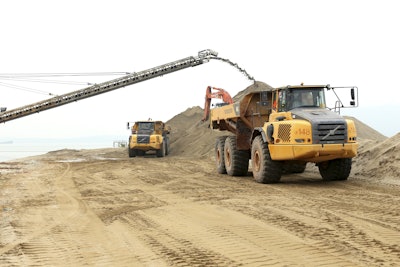 The machines are running tirelessly, loading earth and sand that arrives at the island by barge before being transferred to the fleet of Volvo haulers for delivery to the required deposit points. The trucks travel along an uneven haul road approximately 0.6 miles long, and then unload at the jobsite.