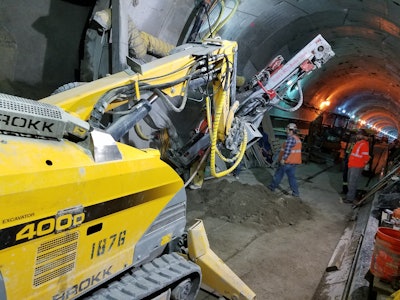 Workers use a Brokk 400D with a TEI 350 rock drill to drill through a tunnel wall. Crews used the attachment to drill holes for steel spiles, well points and freeze pipes.