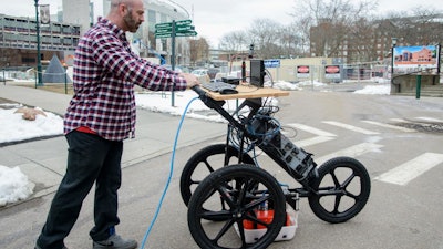 UVM post-doctoral student Dylan Burns demonstrates how the cognitive ground penetrating radar apparatus could be wheeled over an area to scan and map underground infrastructure. The GPR unit, housed in the box near the ground, can peer six to 12 feet down.
