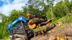 Blue tracked skid steer loader moving large fallen tree logs on rocky terrain with green forest background