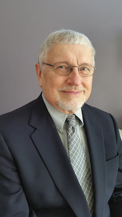 Senior professional in navy suit and glasses with gray beard, smiling