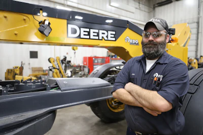 An RDO service technician takes a break from an inspection in front of a John Deere Motor Grader.