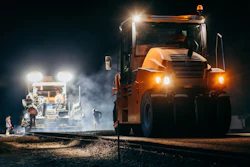 Orange dump truck and asphalt paver operating at night on a road construction site with bright work lights