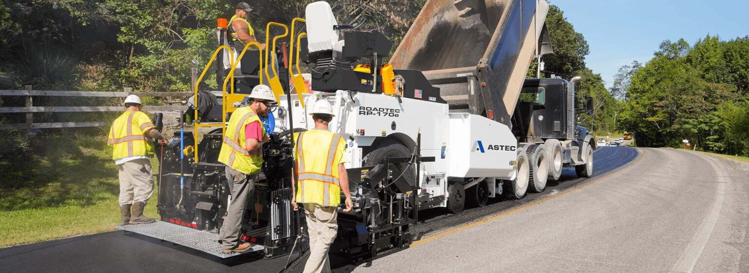 Road construction crew operating asphalt paving equipment on a residential street with safety gear and machinery.