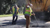 Road construction workers in safety vests operating asphalt paving equipment on a residential street with a paving truck nearby.