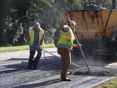 Road construction workers in safety vests operating asphalt paving equipment on a residential street with a paving truck nearby.