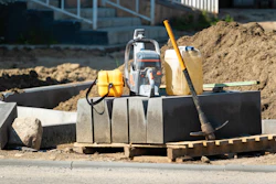 Construction site with compactor, safety vest, fuel container, and tools on concrete foundation
