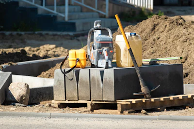 Construction site with compactor, safety vest, fuel container, and tools on concrete foundation