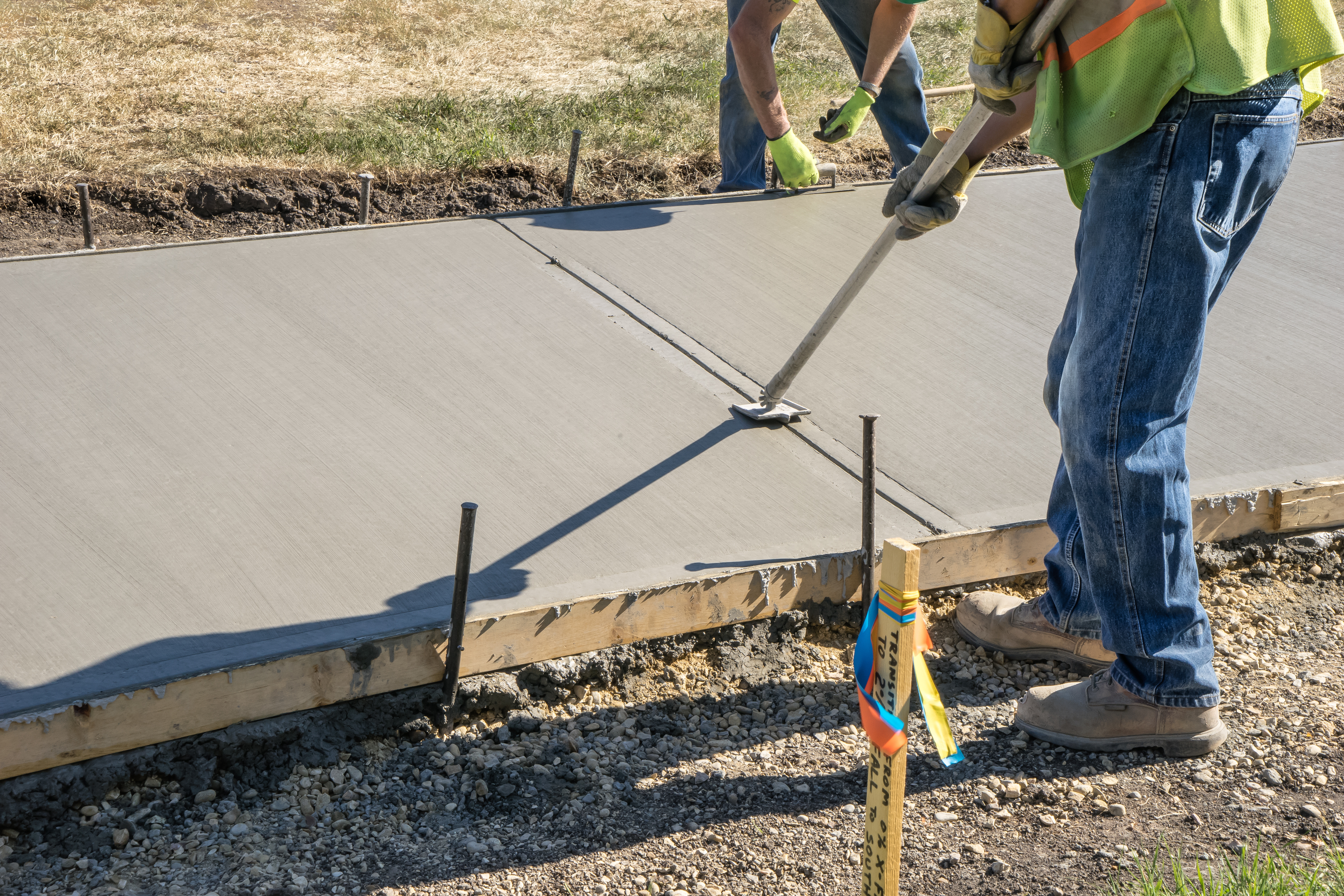 Construction workers finishing wet concrete with hand tools and measurement equipment on a residential driveway project.