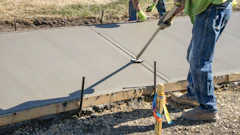 Construction workers finishing wet concrete with hand tools and measurement equipment on a residential driveway project.