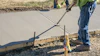 Construction workers finishing wet concrete with hand tools and measurement equipment on a residential driveway project.