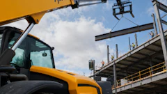 Yellow JCB telehandler crane lifting steel beam at construction site with metal framework