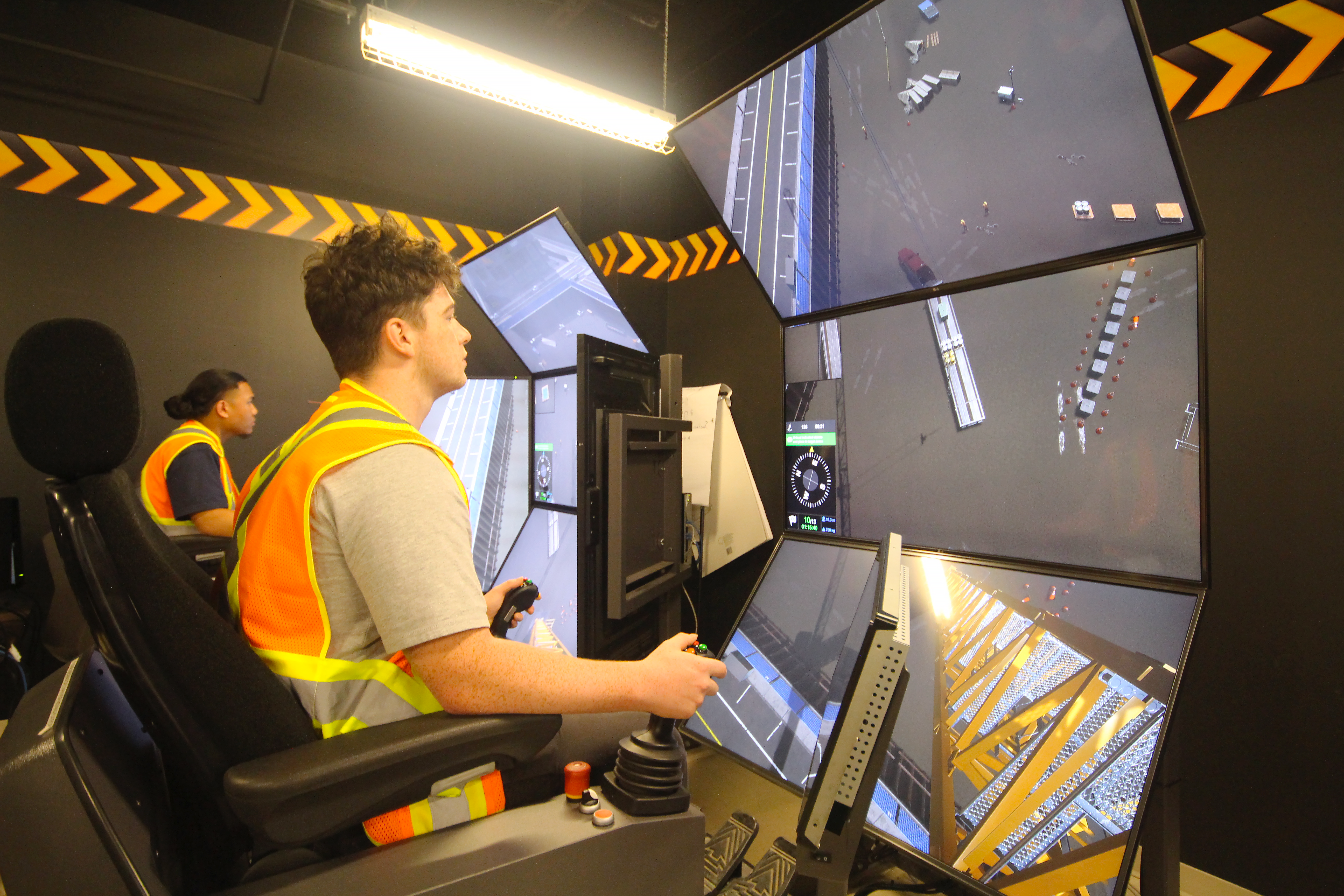 Heavy equipment operator training simulator with multiple monitors and yellow safety striping in control room