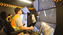 Heavy equipment operator training simulator with multiple monitors and yellow safety striping in control room