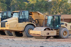 Heavy construction equipment including tan asphalt rollers and compactors lined up at a work site with autumn foliage in background