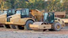 Heavy construction equipment including tan asphalt rollers and compactors lined up at a work site with autumn foliage in background