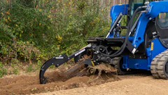 Blue compact track loader with grapple bucket clearing brush and debris from dirt ground near corrugated metal fence
