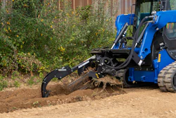 Blue compact track loader with grapple bucket clearing brush and debris from dirt ground near corrugated metal fence