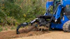 Blue compact track loader with grapple bucket clearing brush and debris from dirt ground near corrugated metal fence