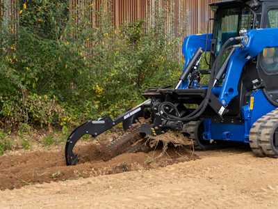 Blue compact track loader with grapple bucket clearing brush and debris from dirt ground near corrugated metal fence