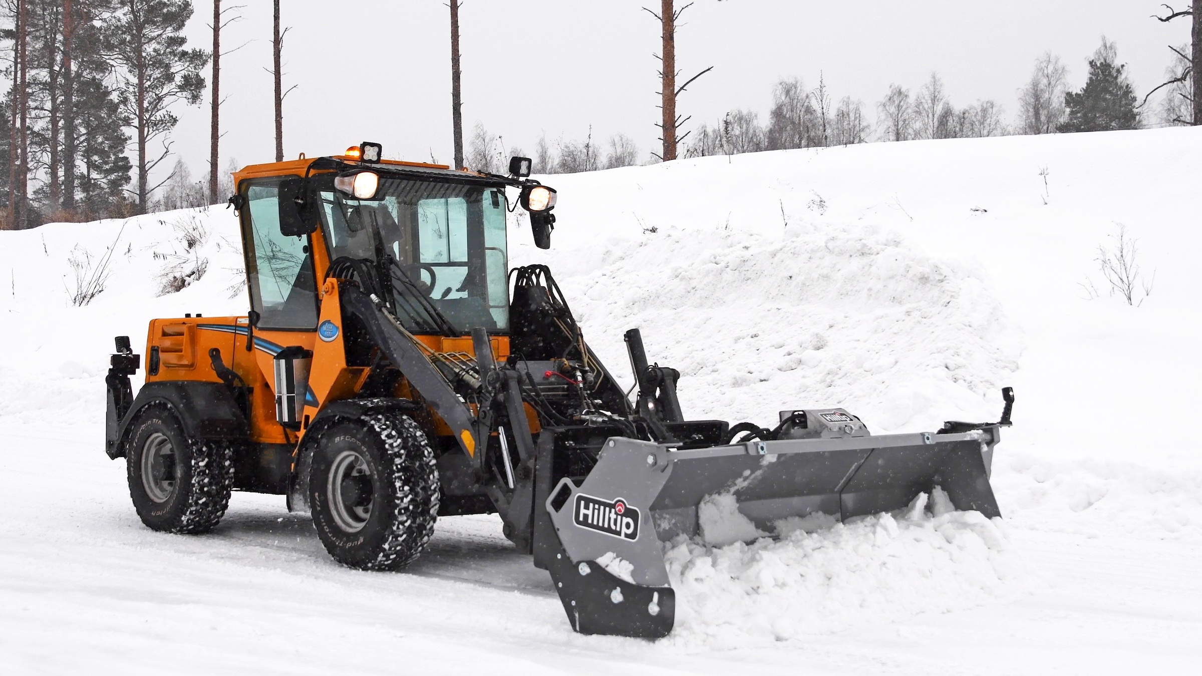Orange wheel loader with Hilltip snow plow clearing snow from a winter landscape