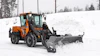 Orange wheel loader with Hilltip snow plow clearing snow from a winter landscape