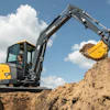 Yellow and black John Deere mini excavator with extended boom digging in sandy soil under blue sky