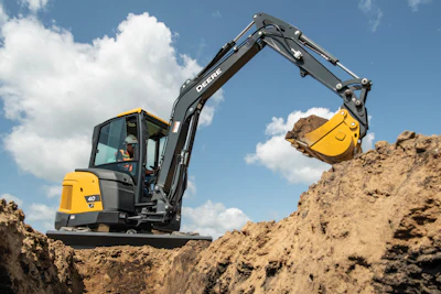 Yellow and black John Deere mini excavator with extended boom digging in sandy soil under blue sky
