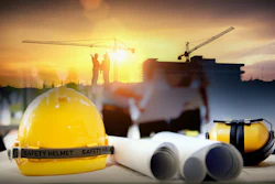 Yellow safety helmet with construction blueprints, ear protection, and workers on building site at sunset
