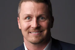Professional headshot of a smiling man in dark blazer and light shirt against gray background