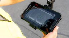 Worker in yellow safety vest holding rugged tablet displaying surveying or mapping software outdoors