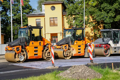 Three orange and black road compactor rollers lined up at a construction site with yellow building in background
