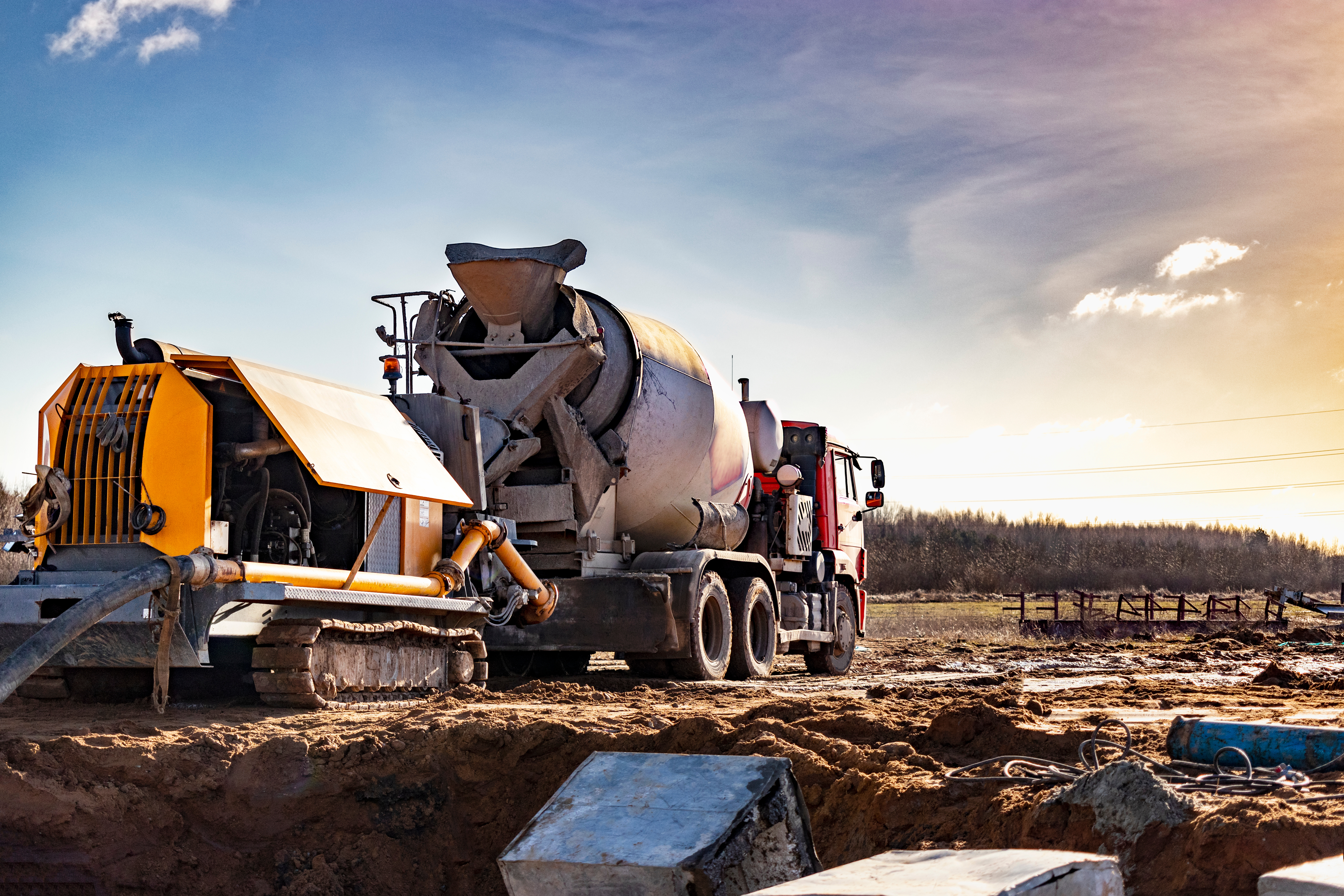 Concrete mixer truck and yellow pump truck operating on construction site with rural landscape
