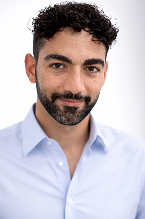 Professional headshot of a man with dark curly hair and beard wearing a light blue button-up shirt against a white background