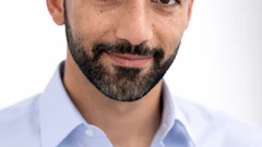 Professional headshot of a man with dark curly hair and beard wearing a light blue button-up shirt against a white background