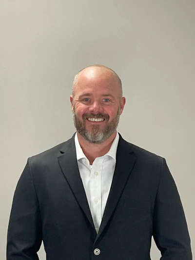 Professional headshot of a smiling man in dark blazer and white shirt against neutral background