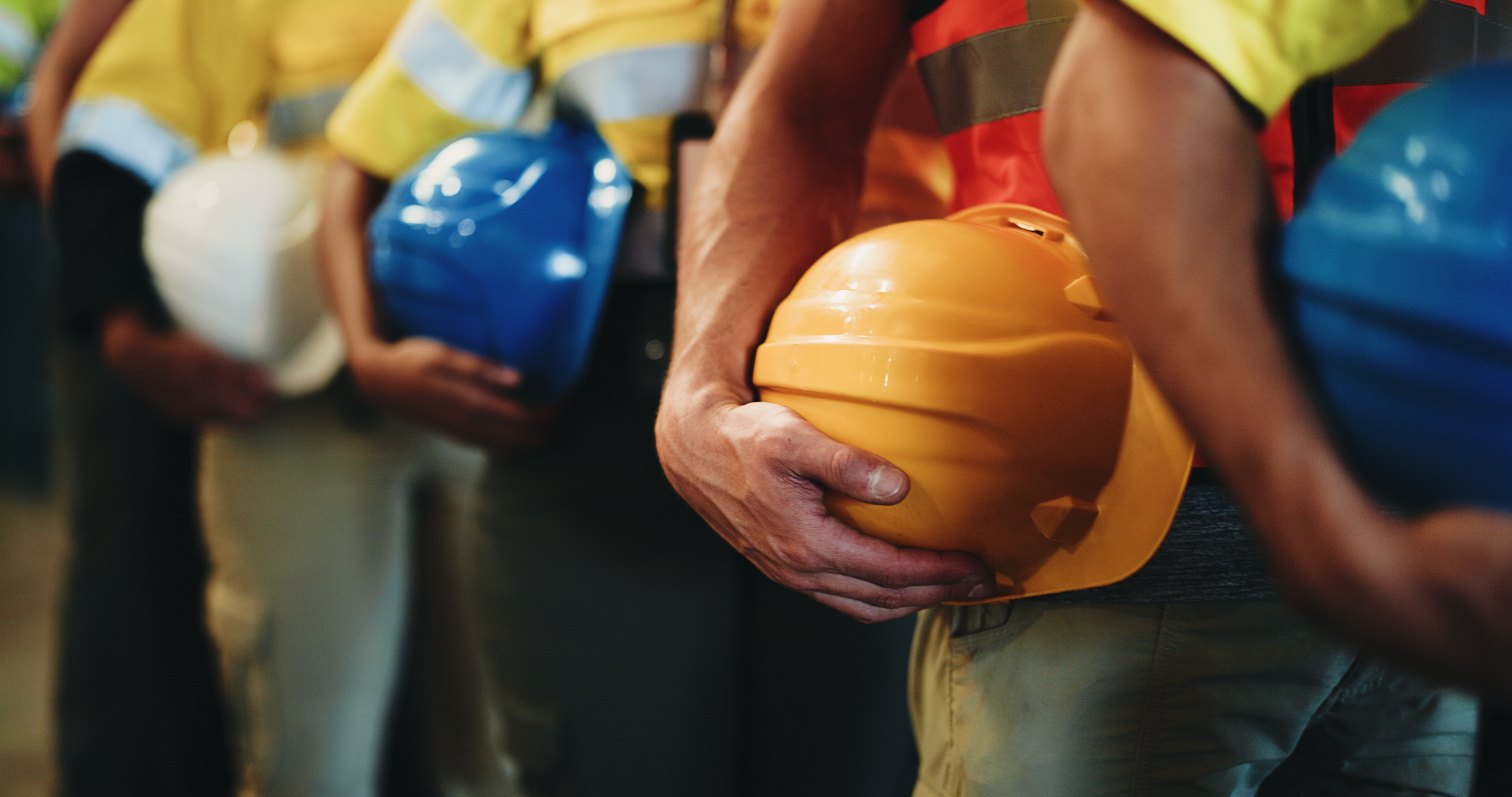 Construction workers in safety vests holding yellow, blue, and white hard hats during a team meeting