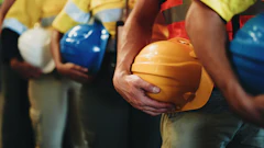 Construction workers in safety vests holding yellow, blue, and white hard hats during a team meeting