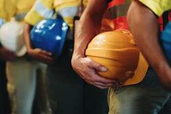 Construction workers in safety vests holding yellow, blue, and white hard hats during a team meeting