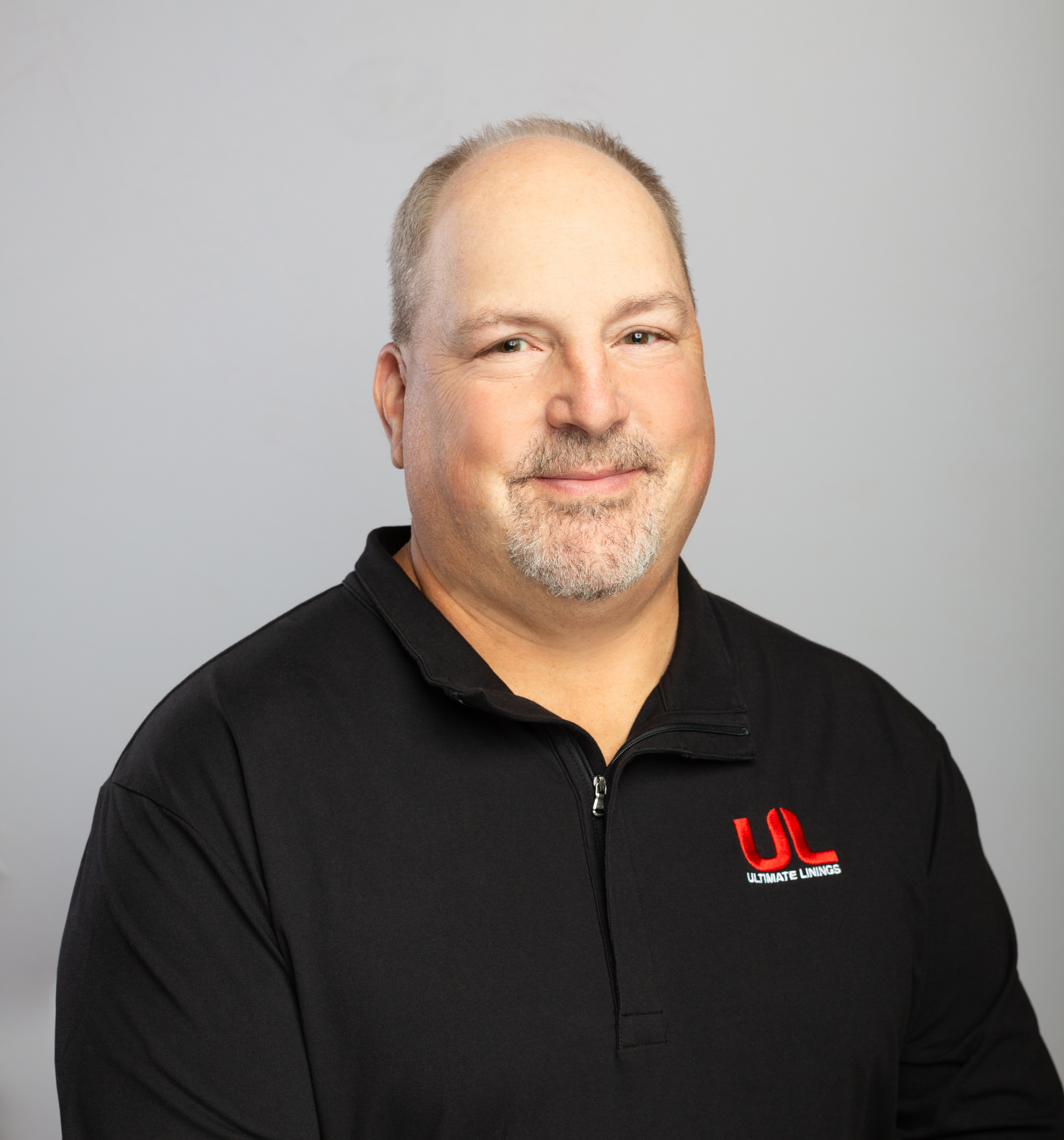 Professional headshot of middle-aged man with goatee wearing black Ultimate Lineman polo shirt against gray background