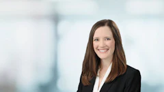 Professional headshot of woman in black blazer and white blouse smiling against blurred office background