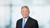 Professional headshot of a man in dark suit and blue tie against blurred office background