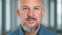 Professional headshot of a middle-aged man with gray hair and beard wearing a blue button-up shirt