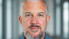 Professional headshot of a middle-aged man with gray hair and beard wearing a blue button-up shirt