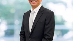 Professional business headshot of an Asian man in black blazer and glasses smiling against blurred cityscape background