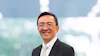 Professional business headshot of an Asian man in black blazer and glasses smiling against blurred cityscape background