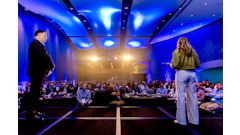 Two business speakers present on stage to an audience in a modern conference hall with blue ambient lighting and warm spotlights.
