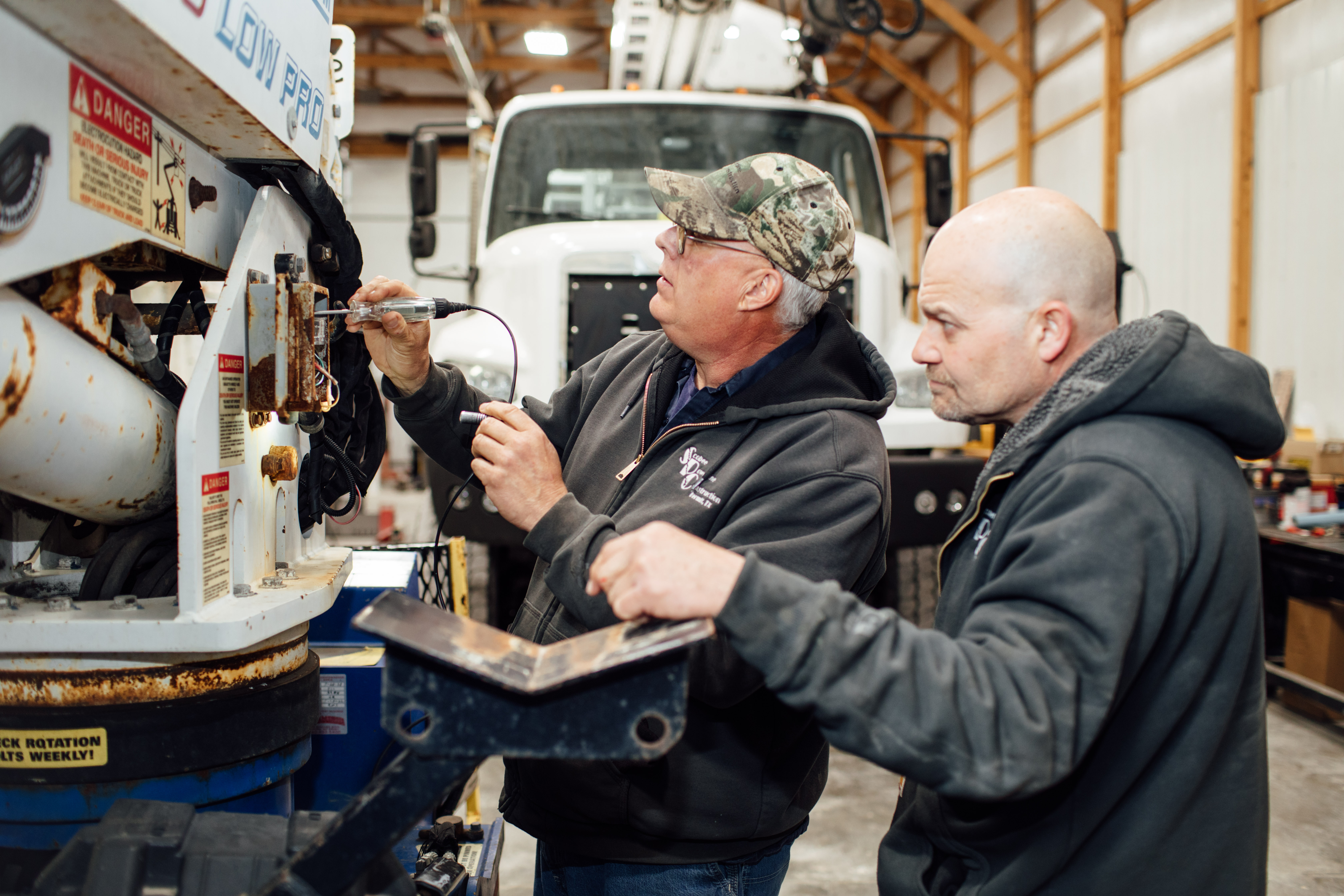 Two workers performing maintenance on industrial equipment in a workshop with a truck visible in the background.